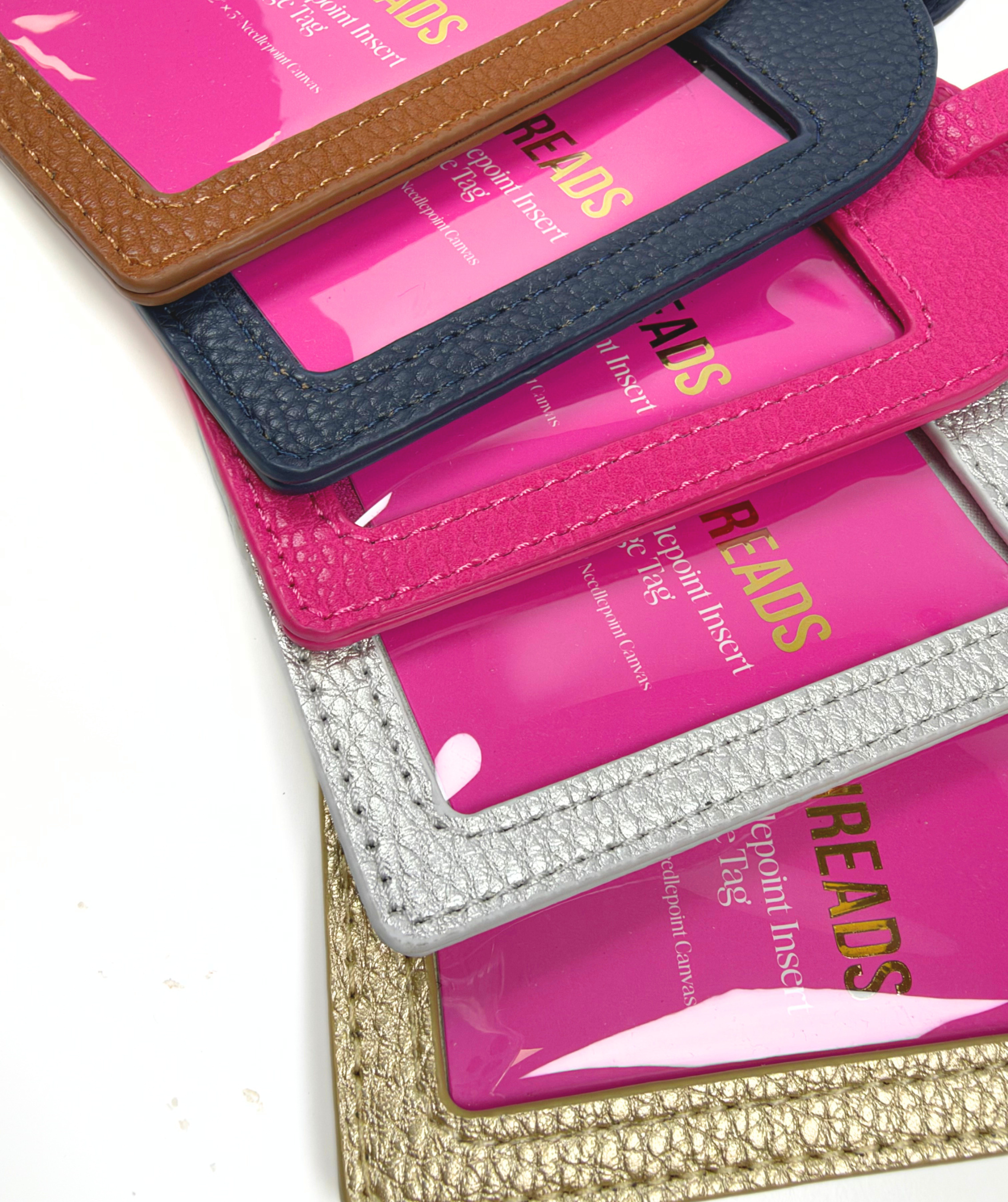 Close-up of Duo Threads self-finishing needlepoint luggage tags in brown, navy, pink, silver, and gold with textured exteriors and clear insert windows.
