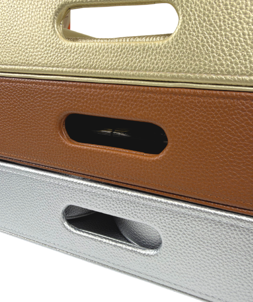 Close-up of stacked self-finishing needlepoint trays in metallic gold, brown, and silver, featuring textured exteriors and built-in cutout handles.
