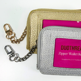 Close-up of gold and silver On-the-Go self-finishing needlepoint wallets featuring textured metallic exteriors, zippered closures, and coordinating clip-on chains.