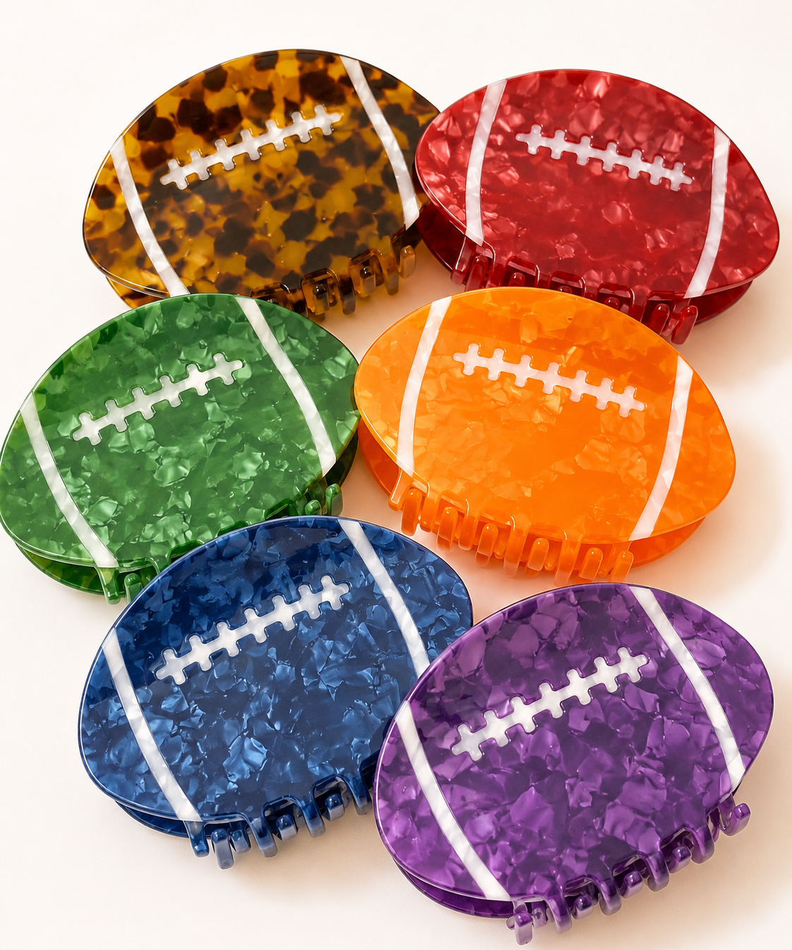 Six football-shaped hair claw clips in assorted colors, including tortoiseshell, red, green, orange, blue, and purple, arranged on a light cream background.