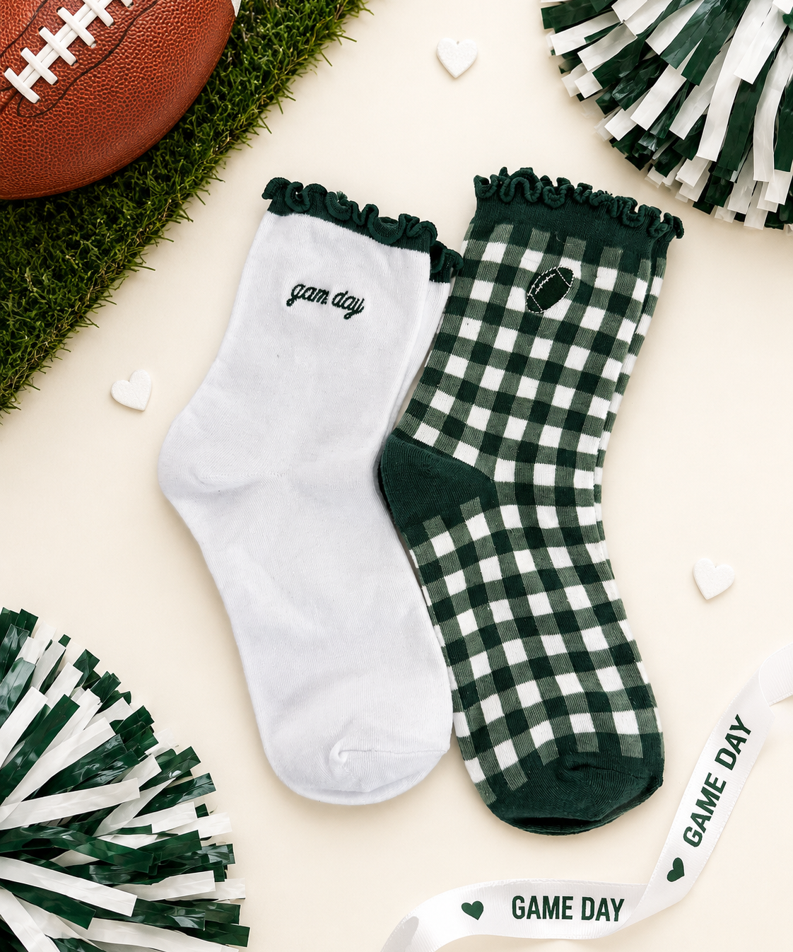 A pair of decorative crew socks laid flat on a cream background with football-themed props. One sock is white with dark green ruffle trim and the word 'game day' in script; the other is dark green and white gingham with a small football patch and 'game day' text.