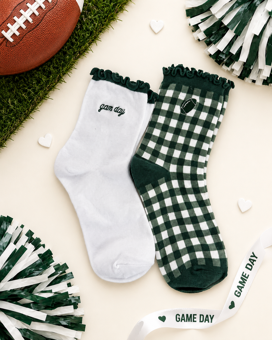 A pair of decorative crew socks laid flat on a cream background with football-themed props. One sock is white with dark green ruffle trim and the word 'game day' in script; the other is dark green and white gingham with a small football patch and 'game day' text.