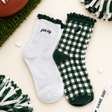 A pair of decorative crew socks laid flat on a cream background with football-themed props. One sock is white with dark green ruffle trim and the word 'game day' in script; the other is dark green and white gingham with a small football patch and 'game day' text.