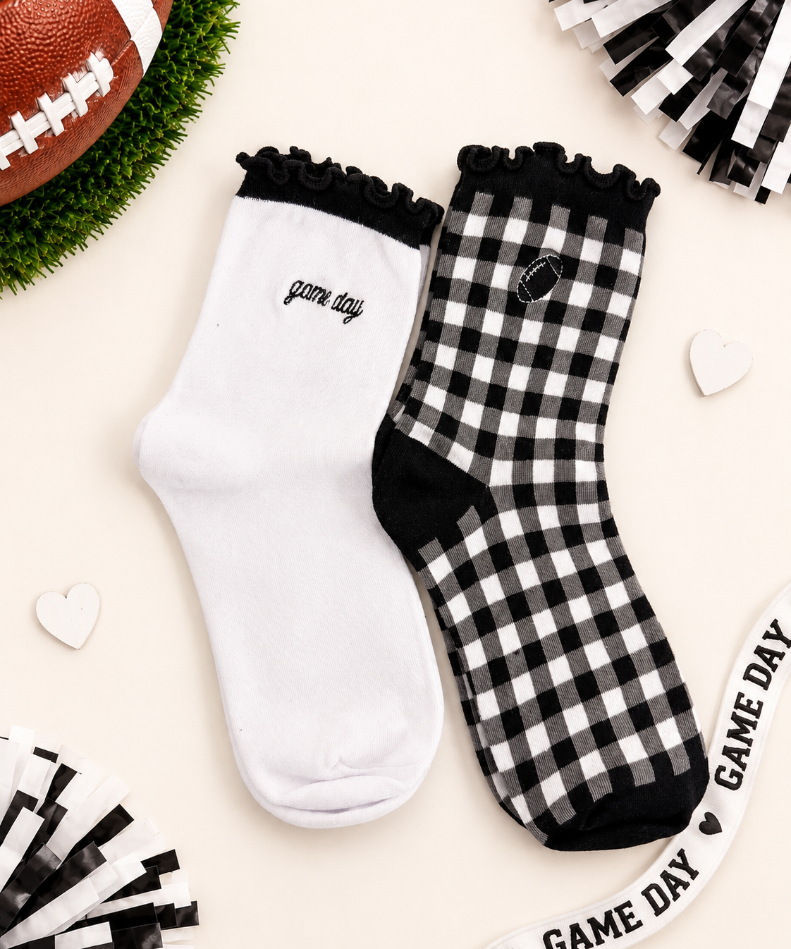 Two socks displayed flat on a light beige background with football-themed props. One sock is solid white with black ruffle trim and 'game day' text, and the other is black-and-white gingham with black ruffle trim, black toe/heel areas, and a small football patch.