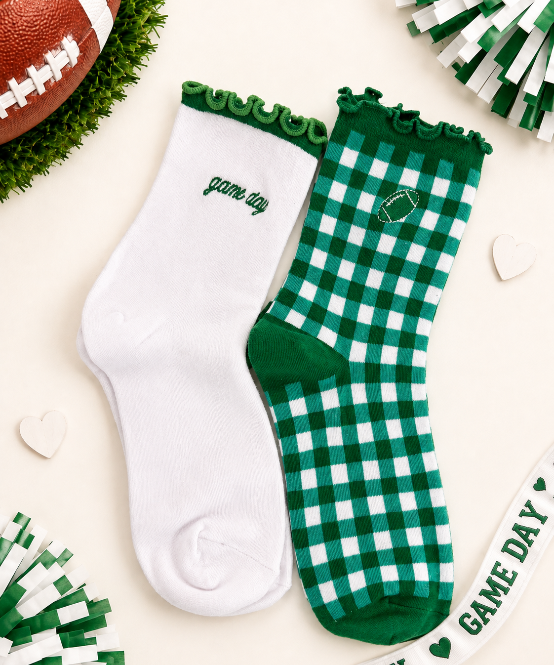 Two socks laid flat on a light cream background with football-themed props. One sock is solid white with green trim and the word 'game day' embroidered near the cuff.