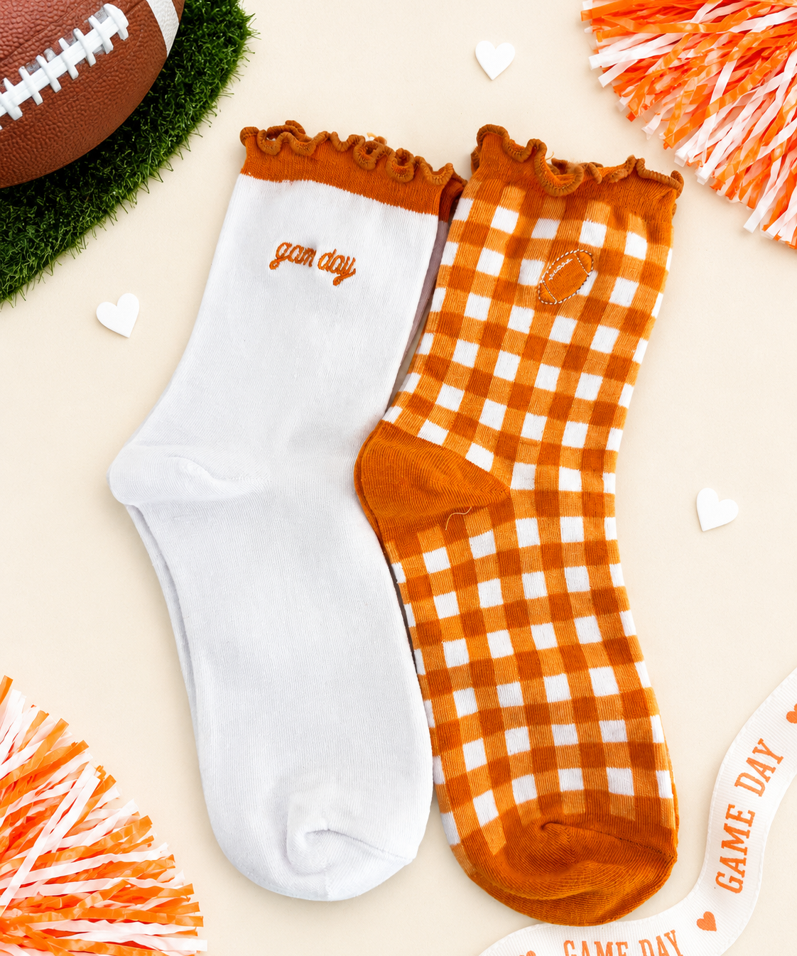 Two crew socks displayed flat on a light beige background with football-themed props. One sock is solid white with orange trim and the word 'game day' embroidered on the side.