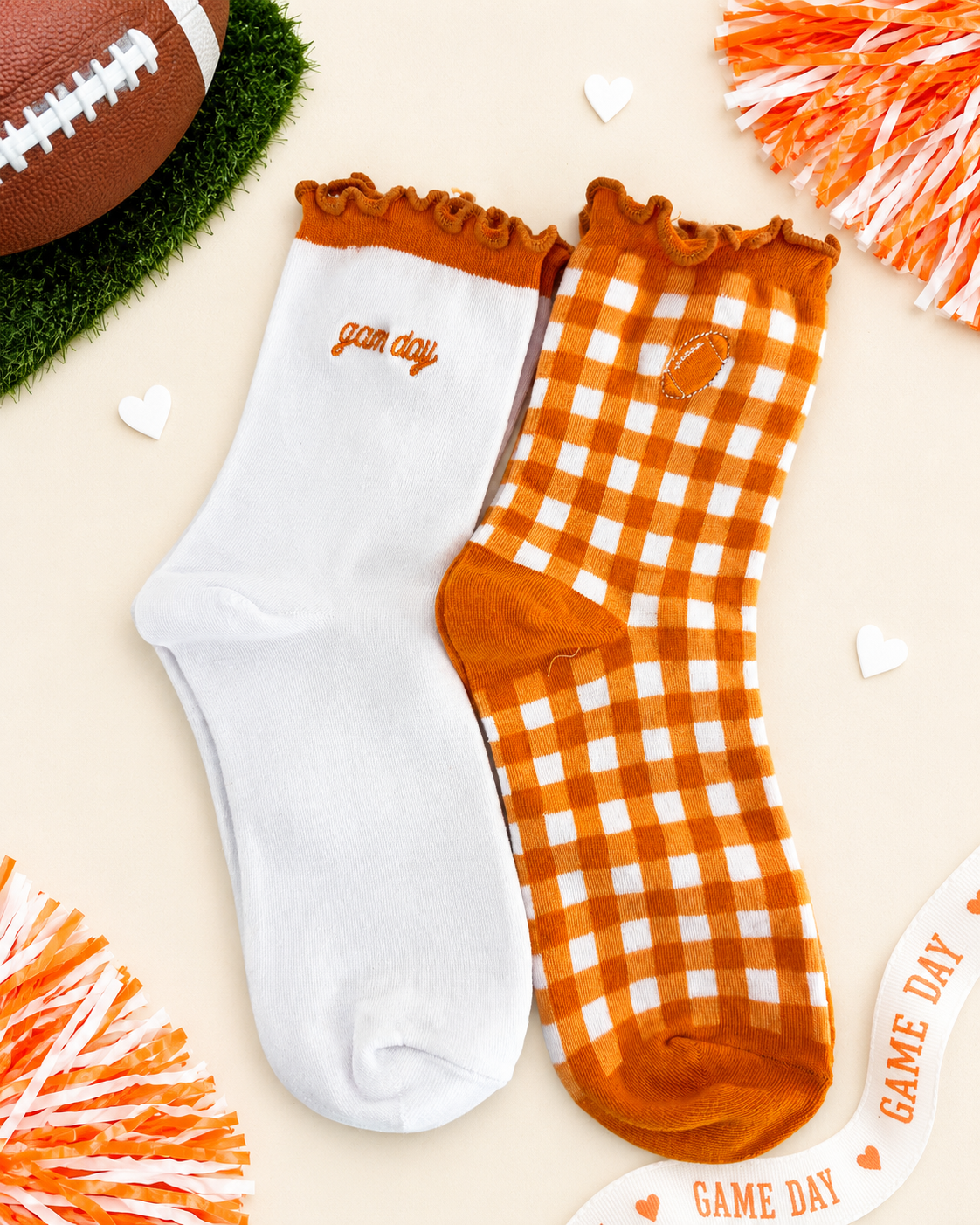 Two crew socks displayed flat on a light beige background with football-themed props. One sock is solid white with orange trim and the word 'game day' embroidered on the side.