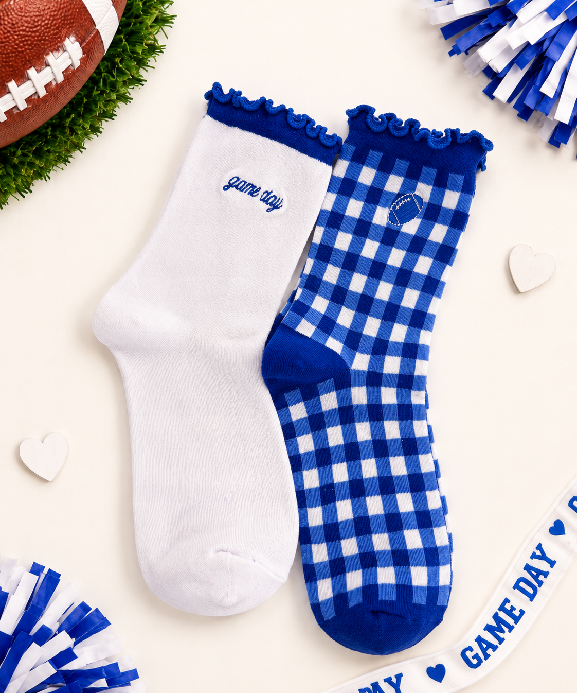Two socks displayed on a cream background with football-themed props. One sock is solid white with royal blue trim and the embroidered text 'game day'.