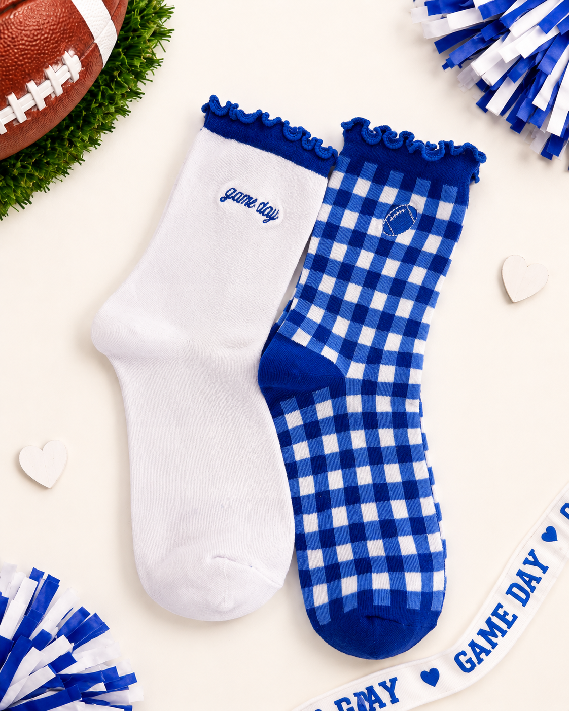 Two socks displayed on a cream background with football-themed props. One sock is solid white with royal blue trim and the embroidered text 'game day'.