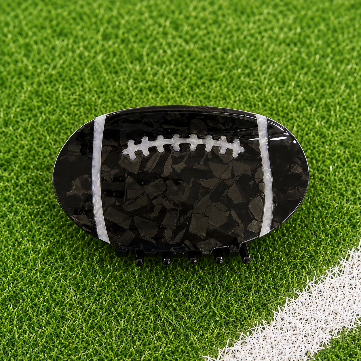A football-shaped hair claw clip with a glossy black and dark gray marbled pattern, white laces, and white side stripes, displayed on a green turf field with a white boundary line.