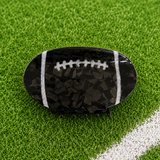 A football-shaped hair claw clip with a glossy black and dark gray marbled pattern, white laces, and white side stripes, displayed on a green turf field with a white boundary line.