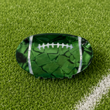 A green football-shaped hair claw clip with a cracked-mercury glass texture and white laces, displayed on a green turf field background.