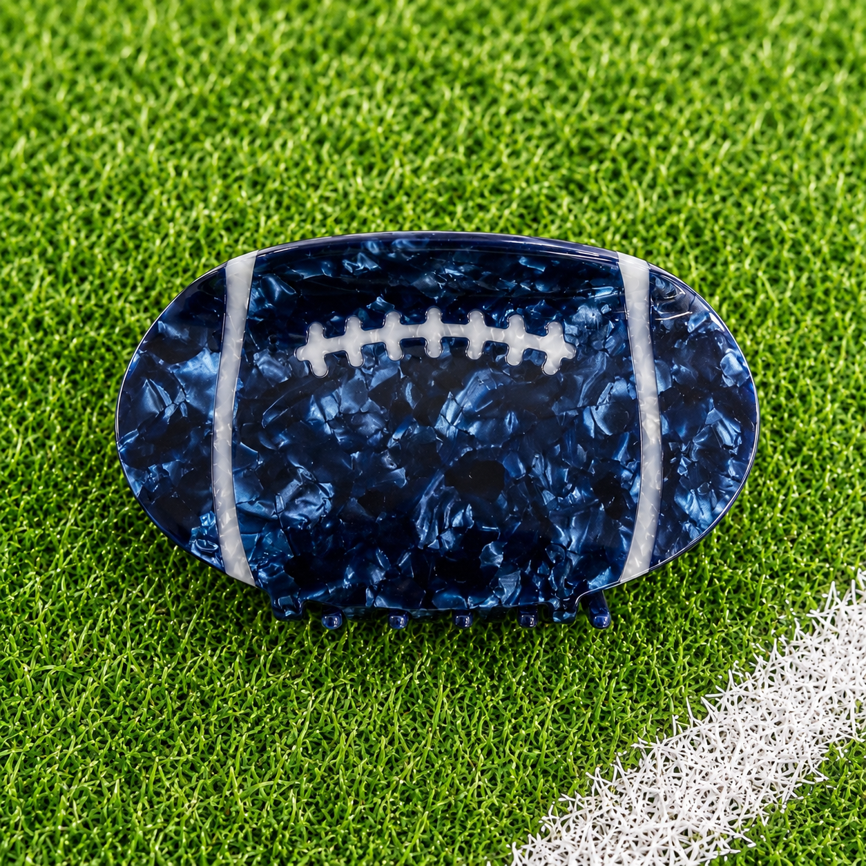 A football-shaped hair claw clip with a dark blue marbled pattern and white laces, displayed on a green grass background.