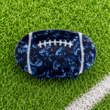 A football-shaped hair claw clip with a dark blue marbled pattern and white laces, displayed on a green grass background.
