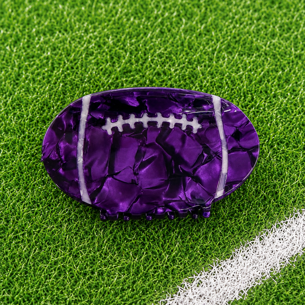 A purple football-shaped accessory with a glossy, cracked metallic texture and white laces, resting on green artificial turf with a white line visible in the corner.