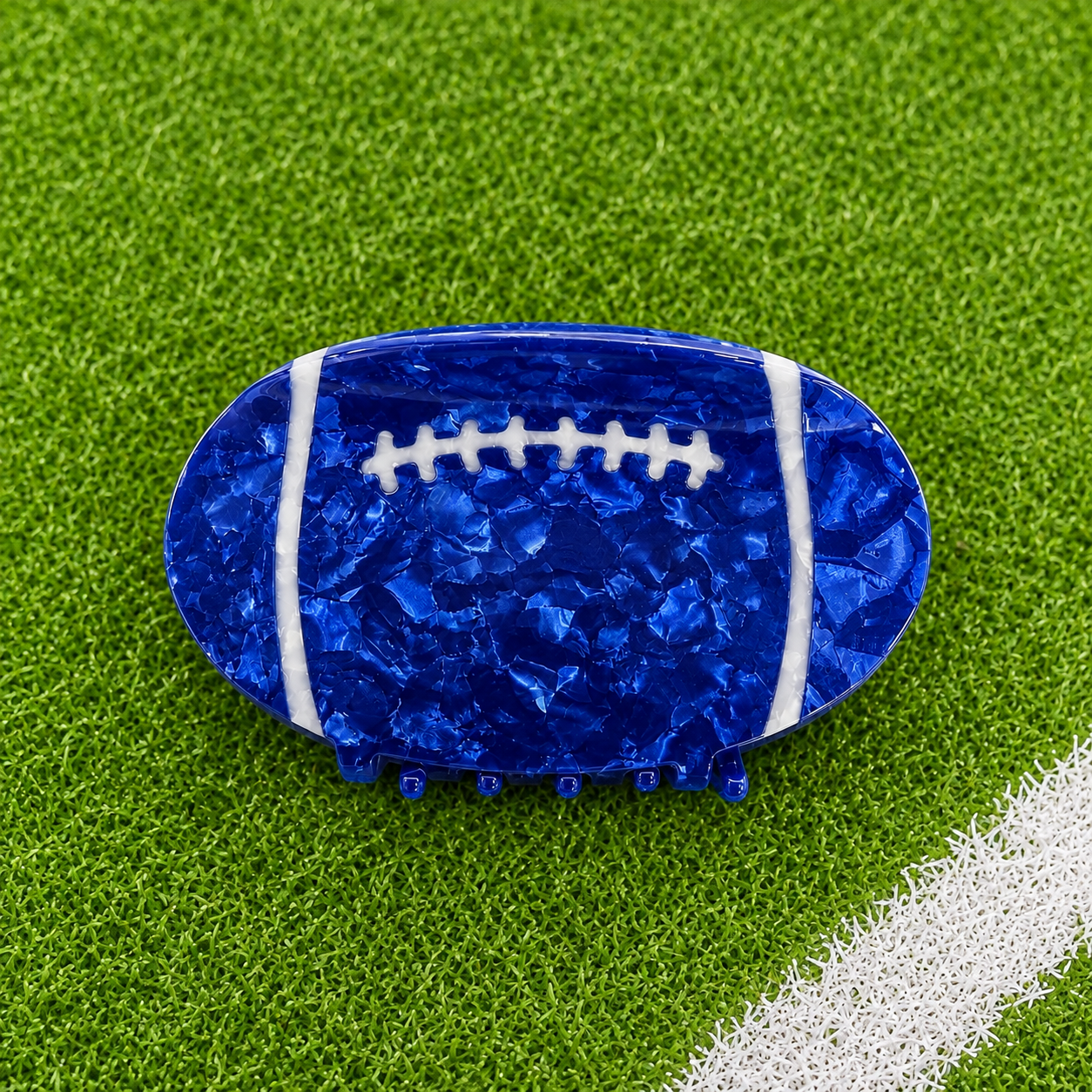 A football-shaped hair claw clip in a glossy, textured royal blue resin with white laces, displayed on a green artificial turf background with a white field line.