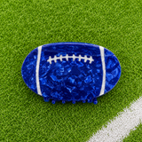 A football-shaped hair claw clip in a glossy, textured royal blue resin with white laces, displayed on a green artificial turf background with a white field line.