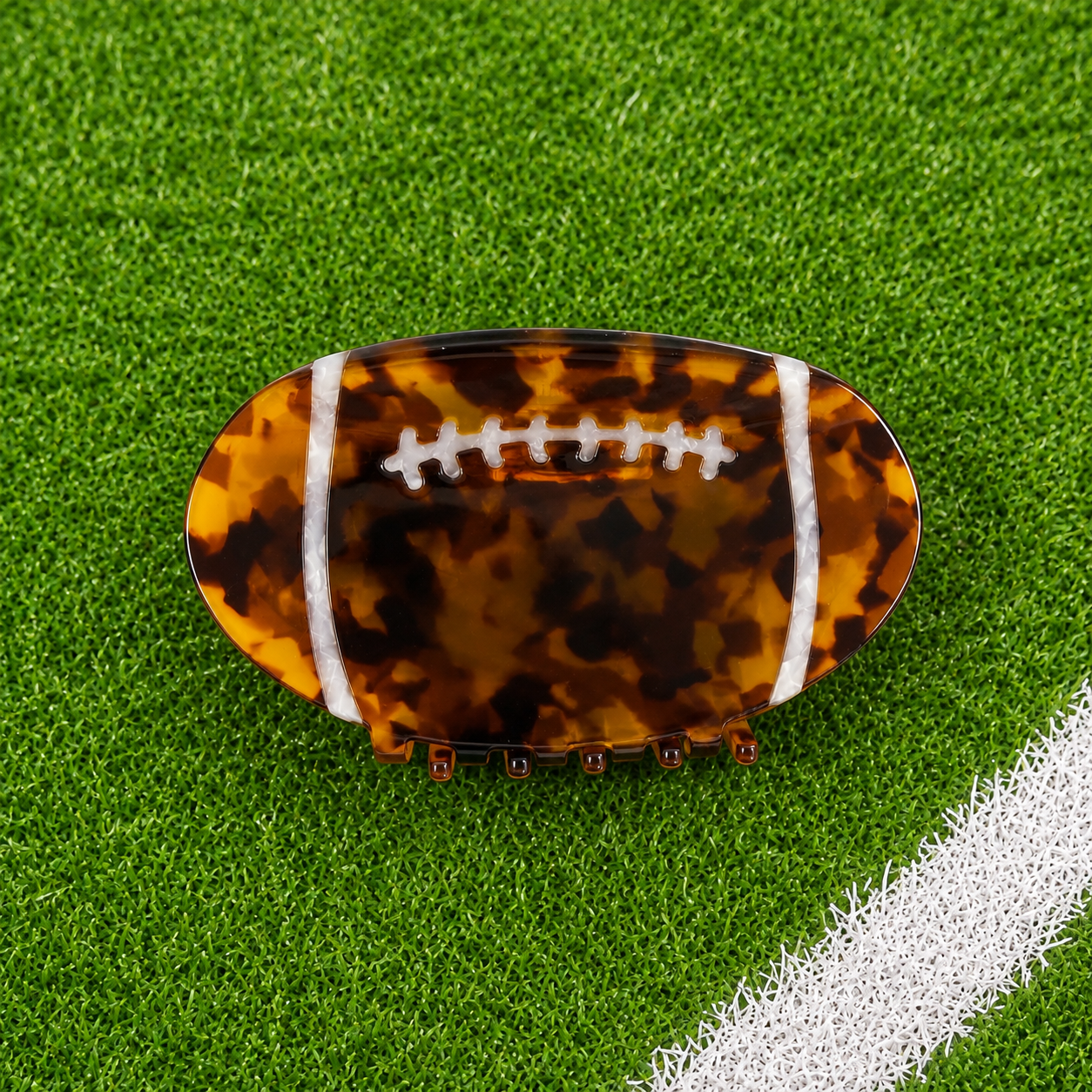 A football-shaped hair claw clip with a brown and black tortoiseshell pattern and white laces, displayed on a green turf background with a white field line.