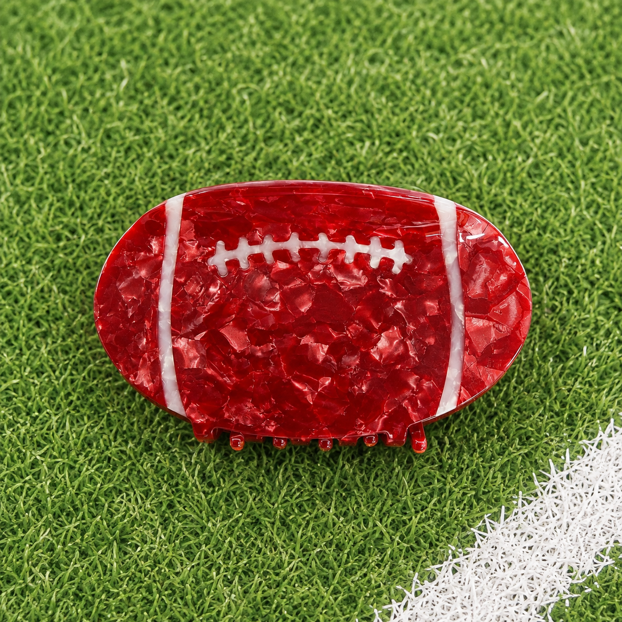 A red football-shaped hair claw clip with a textured, glossy finish and white laces, displayed on a green artificial turf background with a white field line.