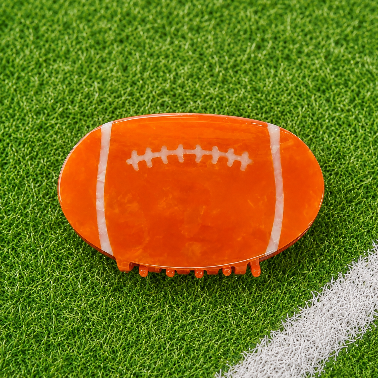 An orange football-shaped hair claw clip with white laces, displayed on a green artificial turf background with a white field line.