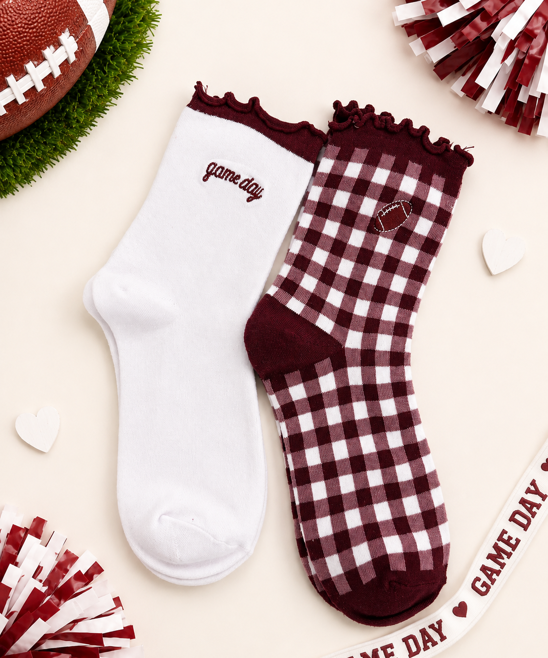 Two ankle socks styled in a flat-lay with football-themed props. One sock is solid white with 'game day' embroidered in burgundy, and the other is a burgundy and white gingham check with a maroon heel, toe, and collar, featuring a small embroidered football patch.