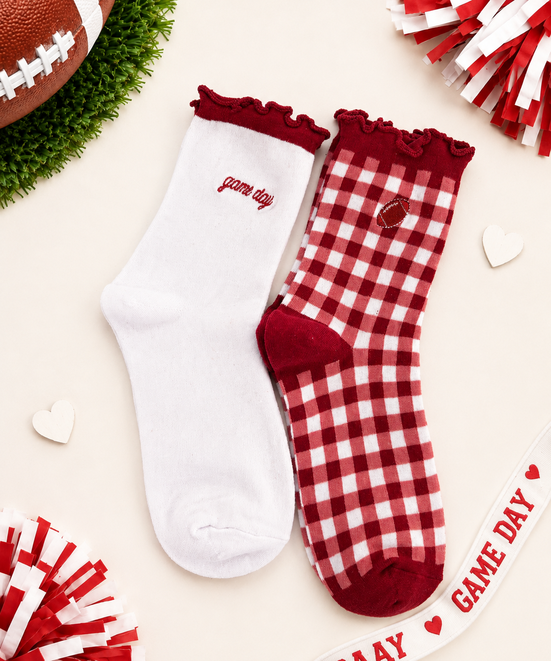 Two decorative ankle socks laid flat on a light cream background with football-themed props. One sock is solid white with scarlet ruffle trim and the word 'game day' embroidered on the side.
