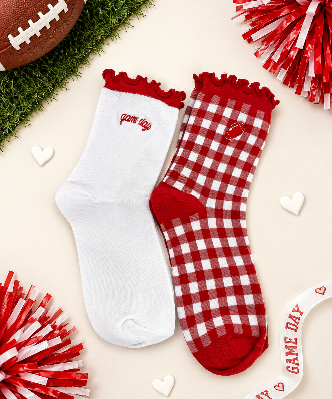 Two red and white socks styled on a light beige background with football and pom-pom props. One sock is solid white with red trim and 'game day' text, and the other is red and white gingham with a small heart patch and red trim.