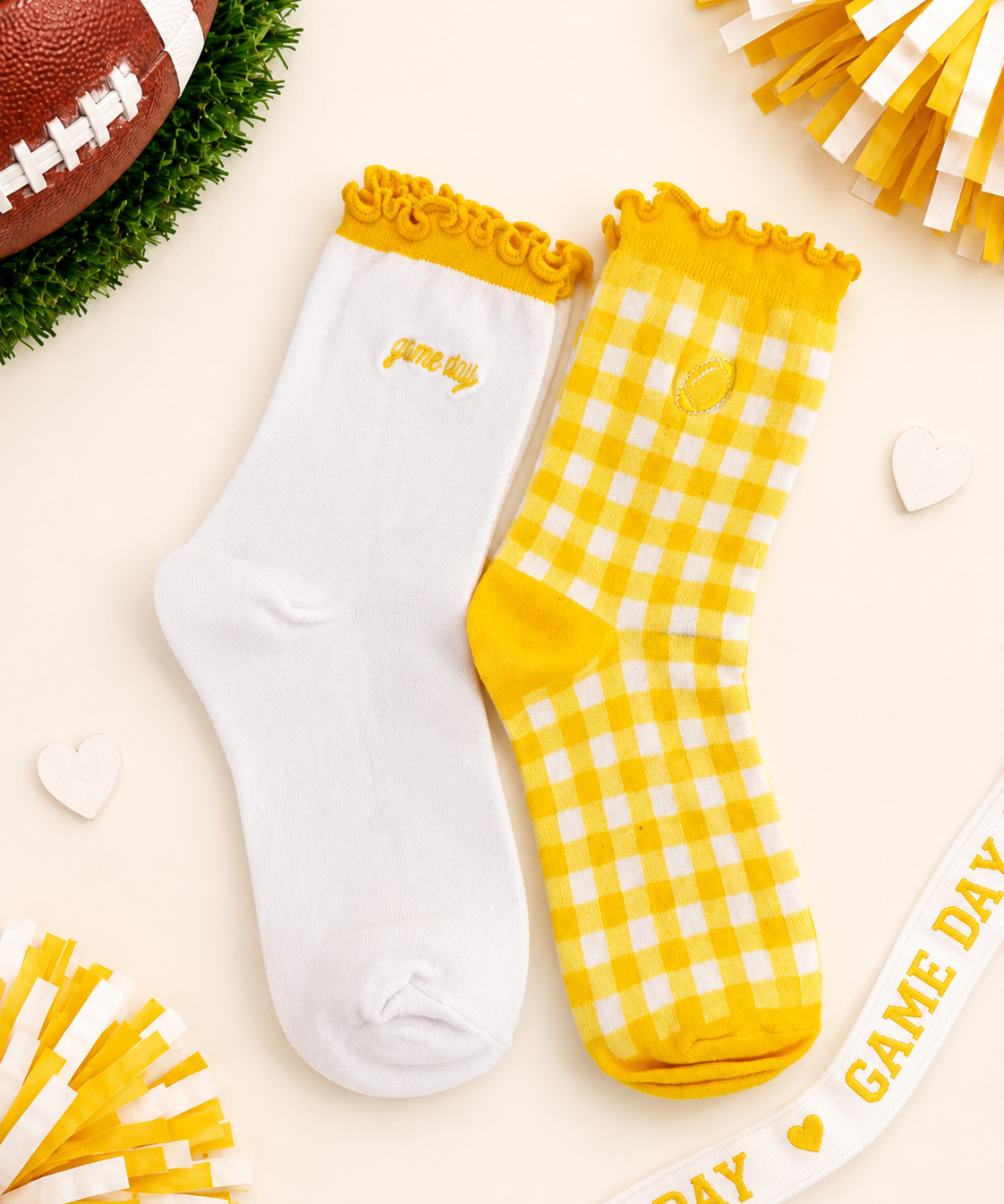 Two socks displayed flat on a light cream background with football-themed props. One sock is white with a yellow ruffled cuff and small yellow 'game day' text; the other is yellow-and-white gingham with a yellow cuff and yellow toe/heel.
